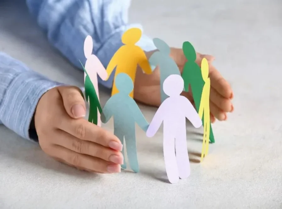 Mãos protegendo um círculo de bonequinhos de papel coloridos, simbolizando inclusão e cuidado com a diversidade